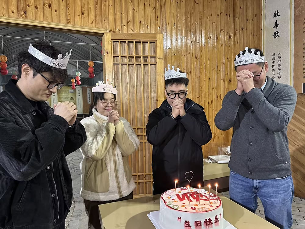 Group of colleagues celebrating Christmas and birthday with candles on cake, wearing festive hats, in a cozy wooden room.